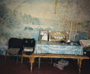In front of faded photo wallpaper is a table with two framed photos, a bench, and two chairs.