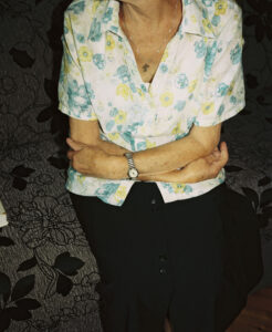 An old woman sits on a sofa, her arms folded.
