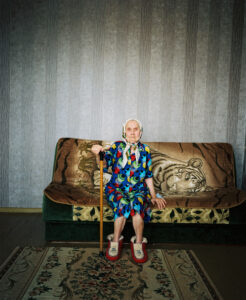 An elderly woman with a walking stick sits on a sofa and looks into the camera.